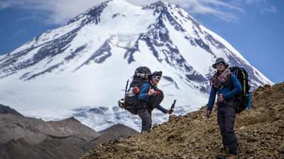 Two men with mountaineering gear stand in front of mountain peak in the Andes.