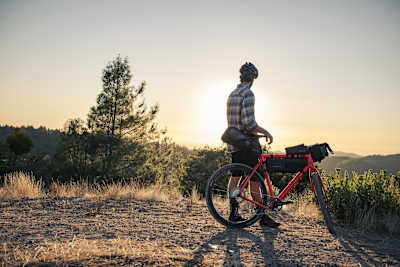 Rider stands with gravel bike looking across mountains and sunset.