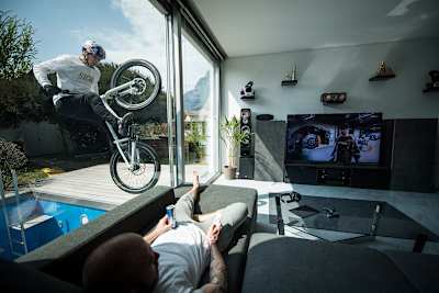 MTB rider Fabio Wibmer rides against glass doors while someone watches TV during filming of his video, Home Office.