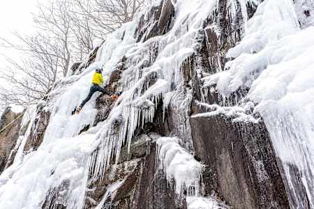 Person in yellow uses ice axe to climb up Frankenstein cliff, New Hampshire.