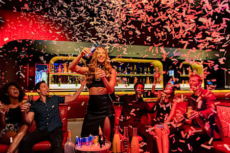 Confetti falling from roof as woman pours herself a Red Bull Energy Drink in front of friends.