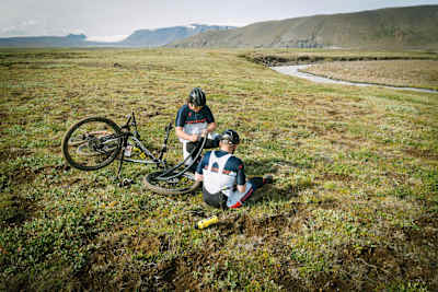 Two MTB riders repair a flat tyre in the wilderness during a ride.