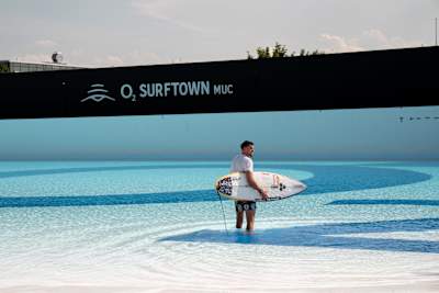 Profi-Surfer Leon Glatzer steht mit seinem Surfbrett unter dem Arm im Wasser des o2 SURFTOWN MUC Wavepools in Hallbergmoos.