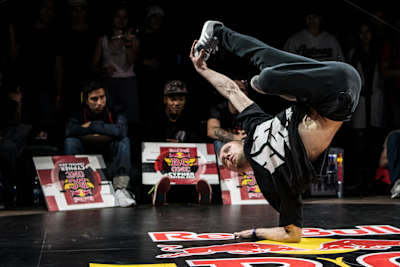 El B-Boy Killa Kolya se sostiene con el codo, en el fondo se puede ver a los jueces.