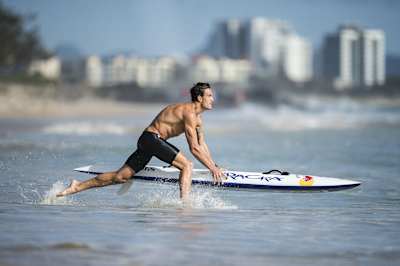 Matt Poole runs out into the sea with his board against a city backdrop.