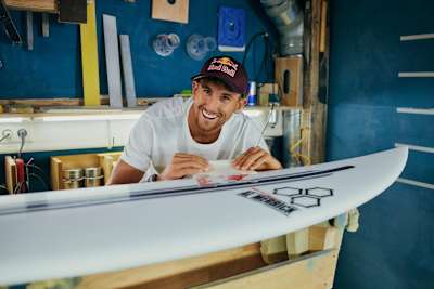 Surf-Profi Leon Glatzer klebt bei einem Foto-Shoot einen Sponsoren-Sticker auf sein Surfbrett.