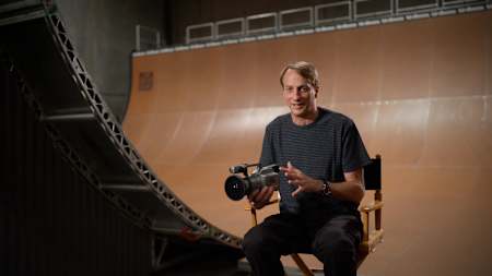 Tony Hawk sitting in front of a skate ramp holding a VX1000 camera.