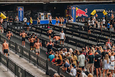 Les spectateurs encouragent les athlètes lors d'une compétition HYROX, tandis que les participants enchaînent les Sandbag Lunges, une épreuve de fentes avec sac lesté.