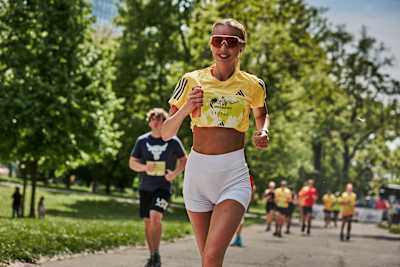 Una sonriente runner con camiseta Adidas amarilla y pantalones cortos blancos participa en Wings for Life, rodeada de otros runners en un camino arbolado.