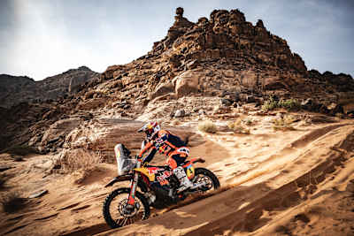 In 2025, Daniel Sanders of KTM Red Bull Factory Racing races his motorcycle across sandy terrain with towering rock formations in the backdrop during Stage 5 of the Dakar Rally in Saudi Arabia