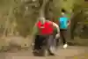 Person in wheelchair on a forest trail with person running behind.