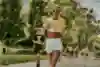 A smiling female runner wearing a yellow Adidas top and white shorts participates in a Wings for Life World Run event, surrounded by other runners on a tree-lined path