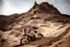 In 2025, Daniel Sanders of KTM Red Bull Factory Racing races his motorcycle across sandy terrain with towering rock formations in the backdrop during Stage 5 of the Dakar Rally in Saudi Arabia