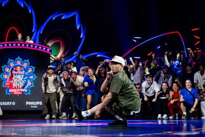 In 2024, MT Pop competes at the Red Bull Dance Your Style World Final event in Mumbai, India, showcasing dynamic street dance moves amidst vibrant lighting and an energetic crowd