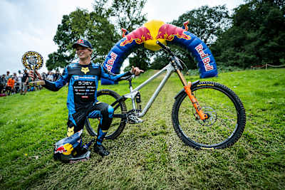 Asa Vermette celebrates winning Red Bull Hardline 2025 in Dinas Mawddwy, Wales, posing with his bike, trophy, and Red Bull helmet beneath the event's iconic arch.