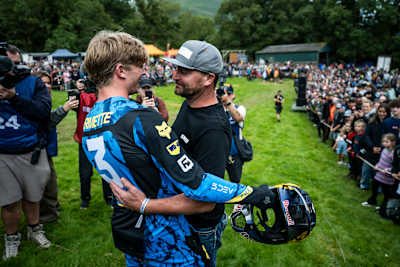 Mountain bike rider Asa Vermette shares a triumphant moment with his father at Red Bull Hardline 2025 in Dinas Mawddwy, Wales, holding his Red Bull-branded helmet before an excited crowd.