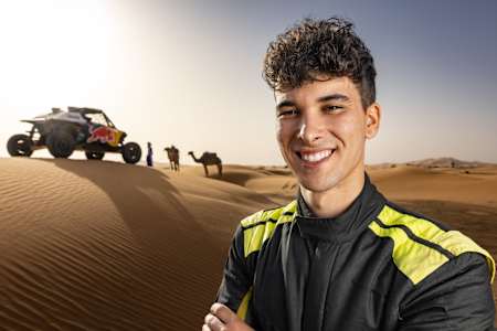 In the Moroccan desert, Gonçalo Guerreiro from Red Bull's Off-Road Junior Team poses in front of a rally car and camels, gearing up for the 2025 Dakar Rally