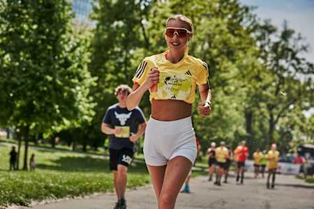 A smiling female runner wearing a yellow Adidas top and white shorts participates in a Wings for Life World Run event, surrounded by other runners on a tree-lined path