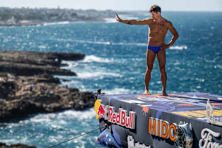 In Italy, 2025, Constantin Popovici stands atop the 27m platform at Red Bull Cliff Diving World Series in Polignano a Mare, embracing the crowd and the dramatic Adriatic coastline