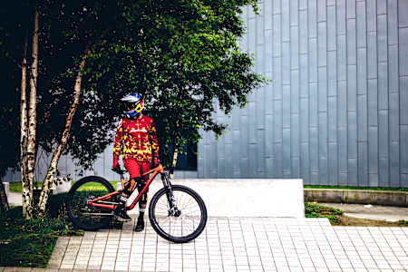 Kriss Kyle poses on a bike in front of a building and next to a tree.