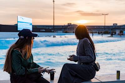 Zwei Frauen sitzen am Rand des o2 SURFTOWN MUC Wavepools und sehen den Surfern in Hallbergmoos beim Surfen zu.