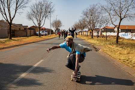 In the foreground, a crouched skateboarder drives towards the camera on a street in Johannesburg. Behind him are his cheering friends, also on skateboards. 