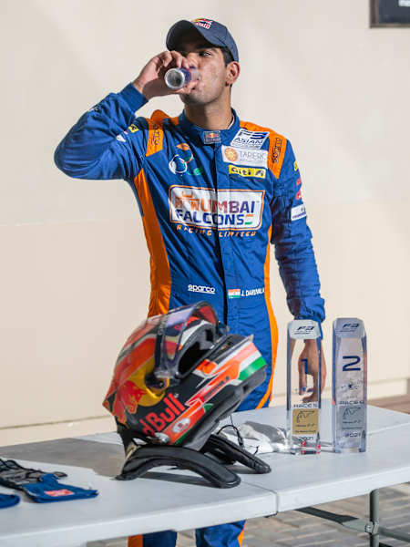 Jehan Daruvala drinks a Red Bull while standing beside his 2021 F3 Asian Championship trophies.