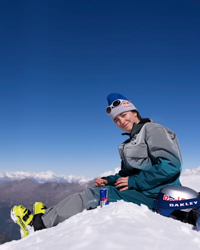 Beim Red Bull Performance Camp 2025 in Saas-Fee, Schweiz, entspannt sich Mathilde Gremaud auf verschneiten Pisten mit Red Bull und Oakley-Ausrüstung unter blauem Himmel.