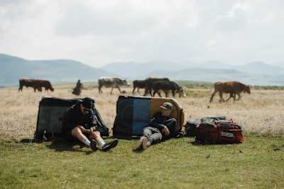 Rob Warner and Matt Jones rest against bike bags on a plain in Lesotho with cows in the background.