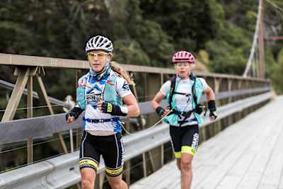 Two women running across a bridge in adventure racing kit.