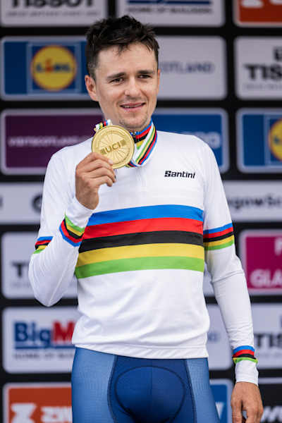 Tom Pidcock proudly holds his gold medal after winning at the 2023 UCI XCO World Championships in Glentress, Scotland, wearing the rainbow-striped champion’s jersey.