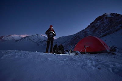 Person stood with head-torch in the snow by tent at the top of the mountain.