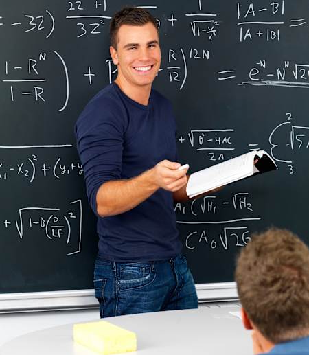 Uśmiechający się student stojący, z książką w ręku, przed tablicą z zapisanymi na niej równaniami.