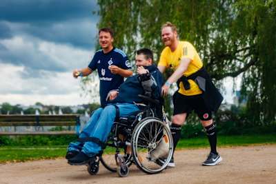 Drei Männer, einer im HSV-Trikot, einer im FC St. Pauli Trikot und einer im Rollstuhl, laufen gemeinsam in Hamburg auf einer Laufstrecke an der Elbe entlang.
