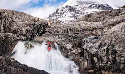 Nouria Newman kayaks off craggy waterfall in Patagonia with snow covered mountain in background.