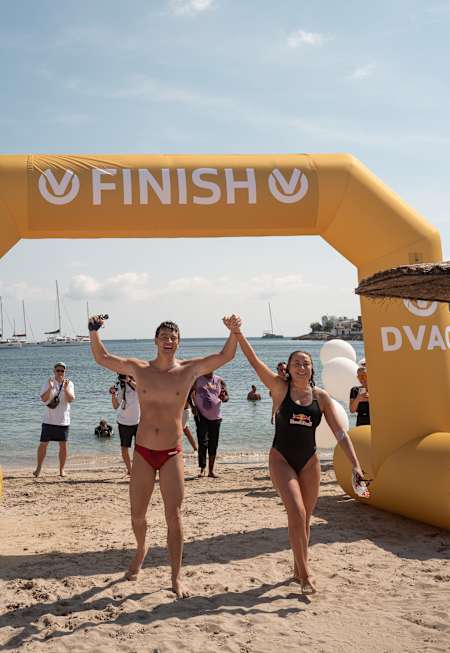 In Ibiza, 2025, Nathalie Pohl and Andy Donaldson triumphantly cross the beach swim finish, raising their hands at the vibrant seaside event.