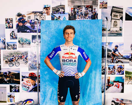 Arne Marit steht stolz im Red Bull - BORA - hansgrohe Trikot für ein Porträt der Saison 2025 in Fuschl, Österreich, mit eindrucksvollen Radsportbildern im Hintergrund.