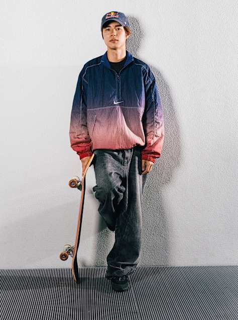 Japanese skateboarder Yuto Horigome leans against a blank wall wearing a Red Bull cap and a Nike jacket, holding his board in his right hand.