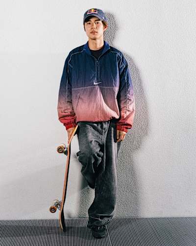 Japanese skateboarder Yuto Horigome leans against a blank wall wearing a Red Bull cap and a Nike jacket, holding his board in his right hand.