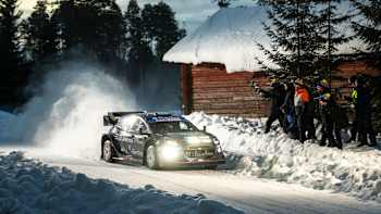 In 2025 WRC Sweden, Jourdan Serderidis powerslides his M-Sport Ford rally car through snowy forest surroundings, kicking up plumes of powder as spectators watch from behind safety barriers