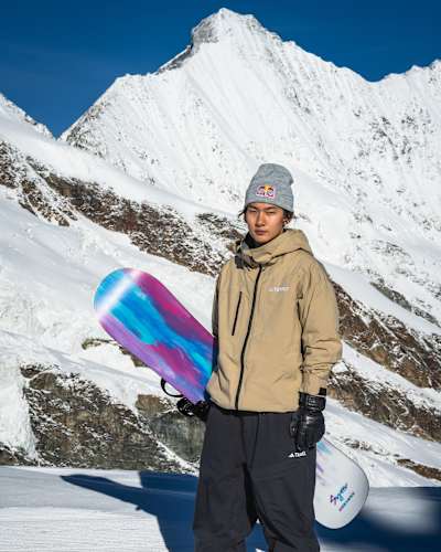 In 2024, Yiming Su poses with a snowboard against snowy mountains at the Red Bull Performance camp in Saas-Fee.