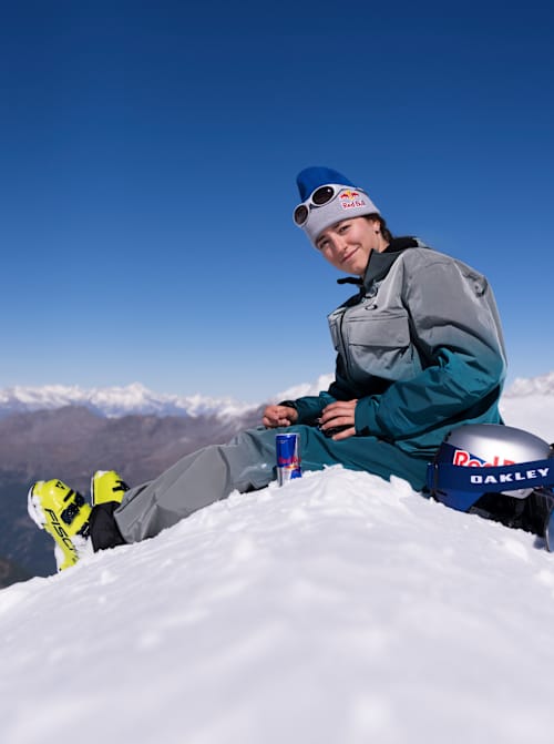 At the 2025 Red Bull Performance Camp in Saas-Fee, Switzerland, Mathilde Gremaud relaxes on snowy slopes with Red Bull and Oakley gear under blue skies.