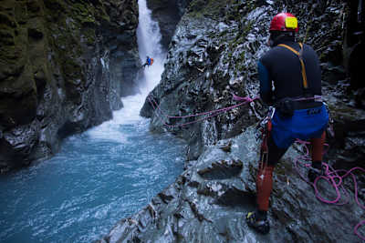 A person abseiling into a canyon is helped by a fellow participant holding rope at Gloomy George, Wanaka, New Zealand.