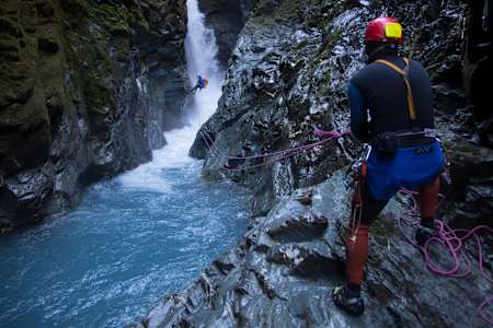 A person abseiling into a canyon is helped by a fellow participant holding rope at Gloomy George, Wanaka, New Zealand.