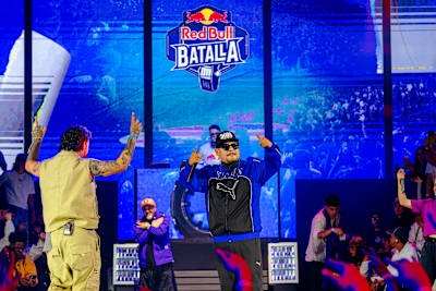Aczino actúa en Red Bull Batalla 2026, animando a un público que abarrota el recinto bajo el logotipo del evento, mientras otros artistas y fans se suman a este vibrante espectáculo de rap en directo