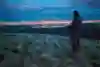 Person stands on hillside with city lights in background.