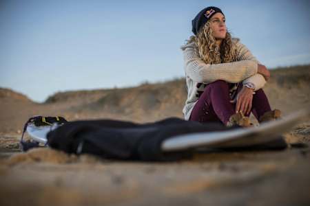 Frankreichs Surf-Ass Justine Dupont sitzt für ein Fotoshooting neben ihrem Surfbrett am Strand und blickt Richtung Meer.