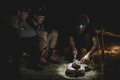 Josh James cuts into a cooked bush animal whille Rob Warner and Matt Jones watch on at their wilderness camp in the Buller District of New Zealand.