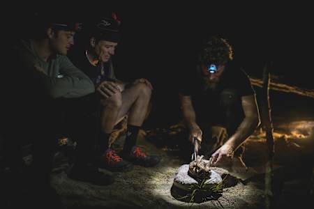 Josh James cuts into a cooked bush animal whille Rob Warner and Matt Jones watch on at their wilderness camp in the Buller District of New Zealand.