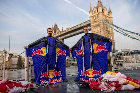 Marco Fürst and Marco Waltenspiel in their Red Bull wingsuits, standing in front of Tower Bridge in London, parachutes lying on the ground beside them